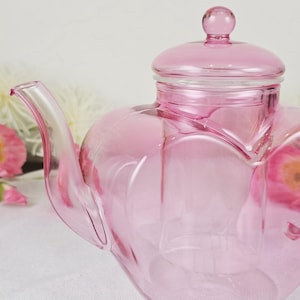 May include: A pink glass teapot with a heart-shaped design. The teapot has a lid with a small knob and a curved spout. The glass is transparent, allowing light to pass through. The teapot is likely used for serving tea.