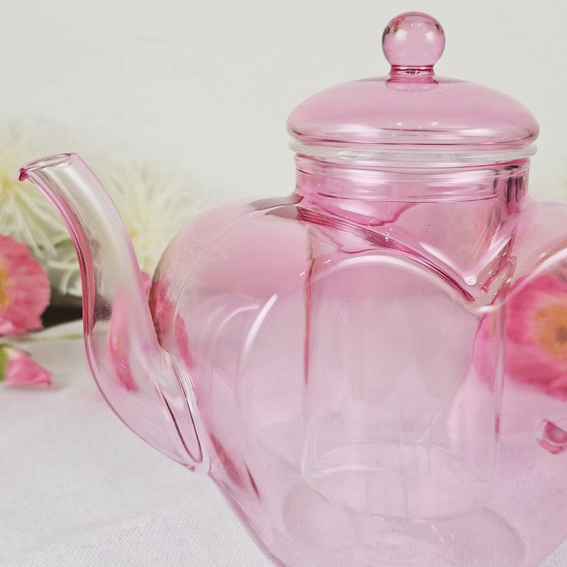 May include: A pink glass teapot with a heart-shaped design. The teapot has a lid with a small knob and a curved spout. The glass is transparent, allowing light to pass through. The teapot is likely used for serving tea.