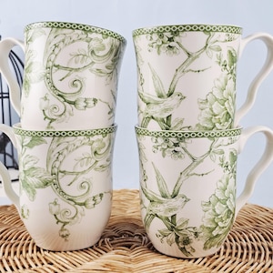 May include: Four white ceramic mugs stacked together. Each mug features a green floral and bird design. The mugs have a curved handle and a green border around the rim. The design includes detailed illustrations of flowers and birds.