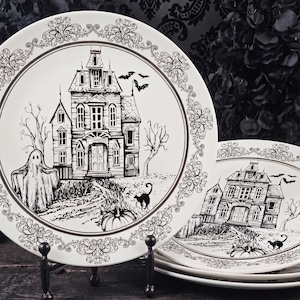 May include: A set of white ceramic plates featuring a black ink illustration of a haunted mansion, a ghost, a black cat, a pumpkin, and flying bats. The design includes an ornate, decorative border pattern surrounding the central scene.