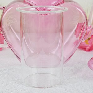 May include: A clear glass double-walled tumbler with a pink tint, set against a soft pink heart-shaped teapot. The tumbler has a wide rim and a cylindrical shape, perfect for serving beverages. The background features a blurred teapot and floral accents.