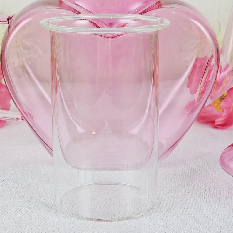 May include: A clear glass double-walled tumbler with a pink tint, set against a soft pink heart-shaped teapot. The tumbler has a wide rim and a cylindrical shape, perfect for serving beverages. The background features a blurred teapot and floral accents.