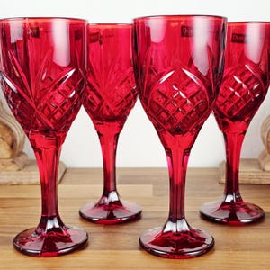 May include: Four ruby red wine glasses with intricate cut glass patterns. The glasses have a tall, slender stem and a wide, rounded bowl. The glasses are arranged on a wooden surface, showcasing their elegant design. The glasses are perfect for serving wine.