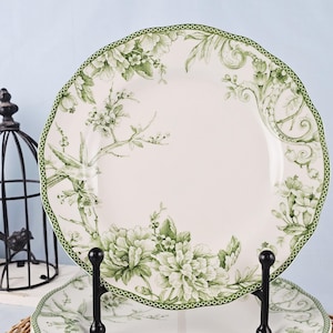 May include: White ceramic plate with a green floral and bird design. The plate has a scalloped edge with a green border. The floral pattern includes peonies and other flowers. The plate is displayed on a black metal stand.