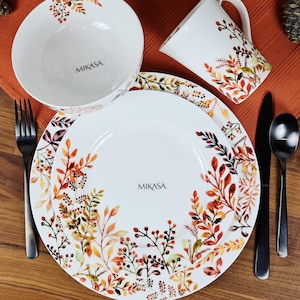 May include: A white Mikasa dinnerware set featuring a bowl, plate, and mug. The set is decorated with a colorful autumn leaf and berry design in shades of orange, red, and green. Includes a fork, knife, and spoon.