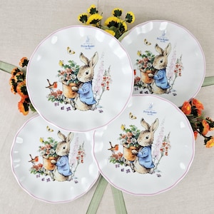 May include: Four white ceramic plates with a scalloped edge and a pink trim. Each plate features a Peter Rabbit illustration with a rabbit holding a flower pot. Butterflies and flowers surround the rabbit. The plates are arranged on a light-colored surface.