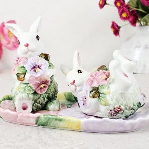 May include: A ceramic Easter bunny figurine set. The white bunnies are adorned with colorful flowers and sit on a pastel-colored tray. The bunnies are surrounded by pink, purple, and green floral accents, creating a charming spring theme.