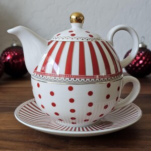 May include: A white ceramic tea set with red accents. The teapot features red stripes and a gold finial. The teacup and saucer have red polka dots and stripes. A festive design for tea time.