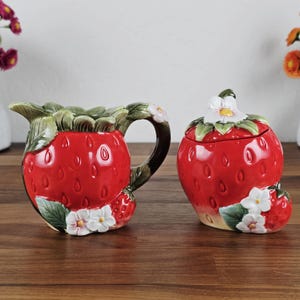 May include: A ceramic strawberry-shaped creamer and sugar bowl set. The creamer has a handle and spout, while the sugar bowl has a lid. Both are red with green leaves and white flower accents. The set is designed to resemble a strawberry.