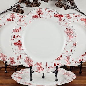 May include: A set of white ceramic plates with a red toile pattern featuring houses, trees, and bridges. The plates have a scalloped edge and a black trim. They are displayed on black metal stands.