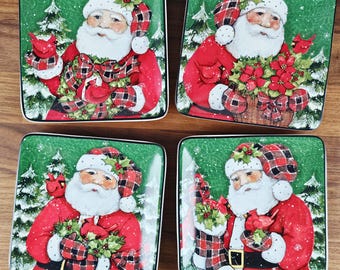 May include: Four square ceramic plates with a Christmas theme. Each plate features a depiction of Santa Claus in a red suit and plaid hat, surrounded by winter foliage and a green background. The plates have a white border.