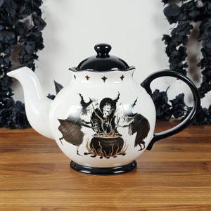 May include: A white ceramic teapot featuring a black lid, handle, and base. The body displays a detailed black and gold illustration of three witches gathered around a bubbling cauldron with flames, accented by small gold fleur-de-lis symbols.