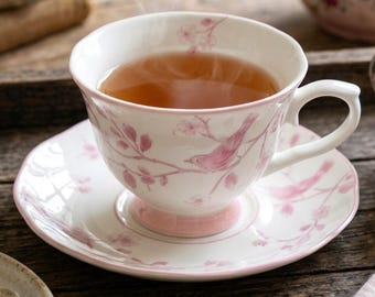 May include: A white porcelain teacup and saucer set featuring a delicate pink pattern of birds perched on flowering branches. The teacup has a curved handle and a pink base, while the saucer has a matching pink rim and floral bird motif.