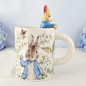 May include: A cream-colored ceramic mug featuring a Peter Rabbit design with a matching rabbit figurine on the rim. The mug has a large, rounded handle and is decorated with floral and butterfly illustrations. The rabbit figurine is wearing a blue jacket.