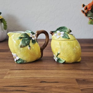 May include: A ceramic lemon-shaped creamer and sugar bowl set. The yellow pieces have a textured surface and are adorned with green leaves and white flowers. The creamer has a brown handle.