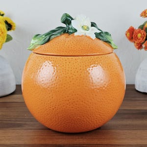 May include: A round ceramic orange-shaped jar with a textured, dimpled orange surface. The lid features a sculpted green leaf and a white blossom with a yellow center. The container has a glossy finish and a spherical body design.
