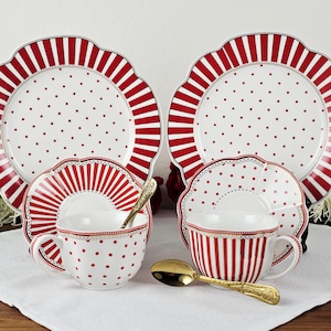 May include: A white and red polka dot and striped tea set. The set includes two plates, two saucers, and two teacups. Gold spoons rest on the white cloth. The plates have a scalloped edge with red stripes.