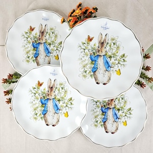 May include: Four white ceramic plates with scalloped edges and gold trim. Each plate features a Peter Rabbit illustration surrounded by white flowers and butterflies. The plates are arranged on a light-colored surface.
