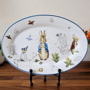 Puede incluir: Plato ovalado para servir con ilustraciones de Peter Rabbit y otros personajes. El plato de cerámica blanca tiene un borde azul y está sostenido por un soporte de metal negro. Las ilustraciones incluyen un conejo con una chaqueta azul y elementos de jardín.