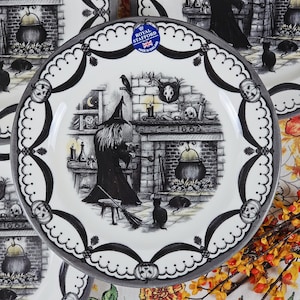 Puede incluir: Un plato decorativo con un diseño de Halloween en blanco y negro. El plato presenta una bruja, un caldero y una escena de chimenea. El borde está decorado con cintas negras y detalles de calabaza. Tiene un sello Royal Stafford.
