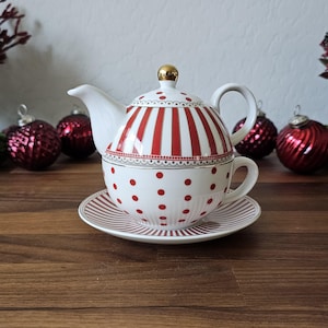 May include: A white ceramic tea set with red stripes and polka dots. The set includes a teapot, teacup, and saucer. The teapot has a gold knob on the lid. The teacup sits inside the teapot. The saucer has red stripes. Christmas ornaments are in the background.