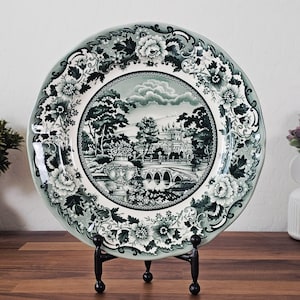 May include: A round ceramic plate featuring a dark green and white transferware pattern. The center depicts a landscape with a stone bridge, a large building, and a decorative urn. The wide rim is adorned with intricate floral and scroll motifs.