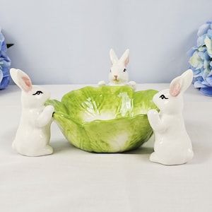 May include: A ceramic Easter centrepiece featuring a green lettuce-shaped bowl with three white bunny figurines. The bunnies have pink-lined ears and are positioned around the bowl, creating a festive and decorative display.