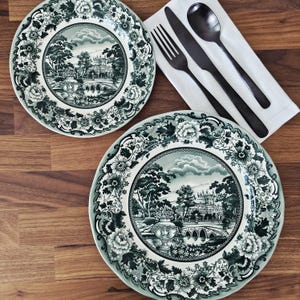 May include: Two green and white decorative plates with a landscape design and floral border. Black cutlery and a white napkin are placed on a wooden surface. The plates feature a bridge and building scene.