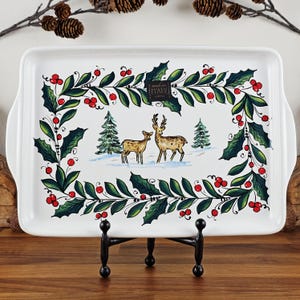 May include: A rectangular white ceramic serving tray with a festive design. The tray features a central winter scene with deer and fir trees, framed by holly and red berries. The handles are curved. The words "made in ITALY" are visible.