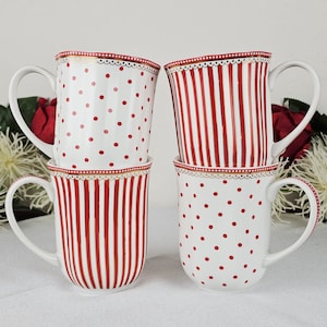 May include: Four white ceramic mugs with red polka dots and stripes. Each mug has a white handle and a decorative gold trim with a scalloped edge. The mugs are stacked together.
