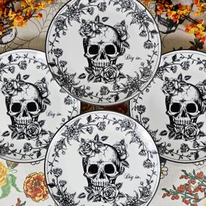 May include: A collection of white ceramic plates with a black and white skull design. Each plate features a skull with a floral crown and the words "Dig in." The plates are decorated with floral patterns.