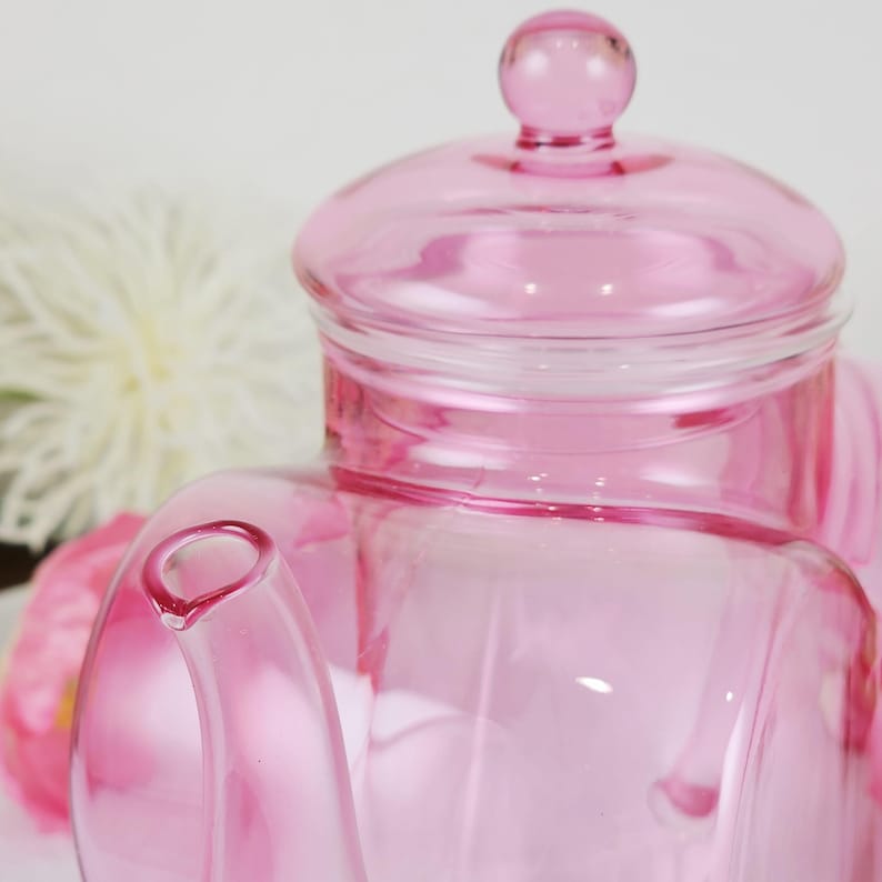 May include: A transparent pink glass teapot with a lid and a small handle. The teapot has a rounded body and a spout. The lid is topped with a small, round knob. The background includes a white flower and pink flowers.