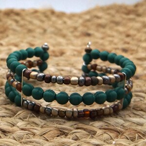 Könnte beinhalten: Ein mehrsträngiges Armband mit dunkelgrünen, braunen und silbernen Perlen. Das Armband hat ein Spiraldesign mit drei Reihen von Perlen. Die Perlen sind auf einem Silberdraht aufgefädelt. Der Hintergrund ist eine gewebte, tanfarbene Oberfläche.