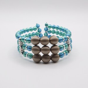 May include: A handmade bracelet featuring three rows of teal and blue beads, interspersed with silver-toned metal accents. The center of the bracelet showcases nine brown, spherical beads. This unique jewelry piece is designed to wrap around the wrist.