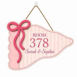 May include: A pennant-shaped wall sign featuring a light pink and white vertical striped pattern. The sign displays the text ROOM 378 and Sarah & Sophia in dark pink. A dark pink bow is attached to the left side, and a brown rope is attached for hanging.