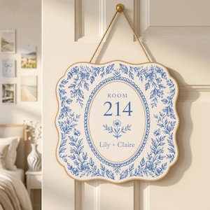 May include: A square wall sign with scalloped edges, featuring a blue and white floral pattern. The center displays an oval frame with the text ROOM 214 and the names Lily + Claire. The sign is suspended by a tan rope from a gold-colored knob.