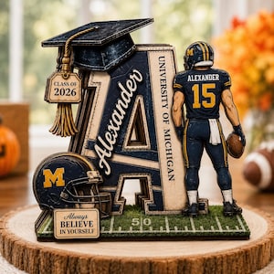 May include: A layered wooden display featuring a large letter A with the name Alexander, a University of Michigan football player figure with number 15, a graduation cap with a Class of 2026 tag, a football helmet, and a sign reading Always Believe In Yourself.
