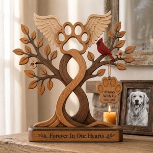May include: A wooden memorial sculpture featuring a central paw print with angel wings, flanked by tree branches with leaves and flowers. A red cardinal sits on a branch, and a paw-shaped tag reads Always With Us 1976-2026. Forever In Our Hearts.