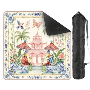 May include: A square picnic blanket featuring a chinoiserie-inspired print with monkeys, a pagoda, butterflies, and floral motifs in blue, pink, and cream tones. The blanket has a black underside and comes with a black cylindrical carrying bag.