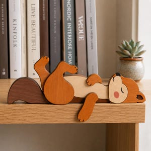 May include: A wooden fox figurine in a reclining position with its eyes closed. The fox features a dark brown tail, a light tan belly, and a reddish-brown head and limbs. It has small black painted eyes and pink circular cheeks on its face.