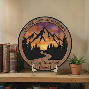 May include: A round wooden plaque with a mountain silhouette design and the text "The best views are yet to come." The plaque has a sunset background and the words "Happy Retirement [Name]" at the bottom.