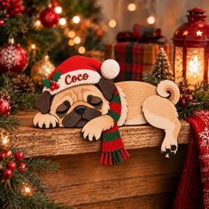 Könnte beinhalten: Ein Holzausschnitt eines schlafenden Mopses, der eine rote Weihnachtsmütze mit dem in Weiß gestickten Namen Coco und einen rot-grün gestreiften Schal trägt. Der Mops hat beiges Fell, eine schwarze Schnauze und ruht mit dem Kopf auf seinen Pfoten.