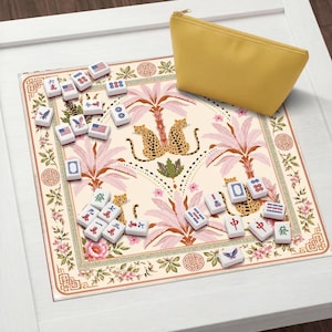 Op de afbeelding: Een mahjongset met witte tegels voorzien van diverse symbolen, cijfers en Amerikaanse vlaggen, gerangschikt op een rechthoekige mat met een crèmekleurige achtergrond, roze palmbomen en luipaardillustraties, inclusief een effen geel etui.