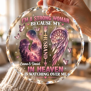 Peut inclure: Une décoration en verre ronde et facettée avec le texte I'M A STRONG WOMAN BECAUSE MY HUSBAND IN HEAVEN IS WATCHING OVER ME. Elle présente la silhouette d'un couple, une grande aile d'ange, des papillons et les noms Emma & Daniel.
