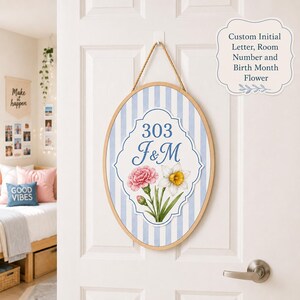 Könnte beinhalten: Ein ovales Wandbehang aus Holz mit einem hellblau-weiß gestreiften Hintergrund. In der Mitte befindet sich ein weißes, gewelltes Schild mit der Nummer 303, den Initialen F&M sowie Illustrationen einer rosa Nelke und einer weißen Narzisse.