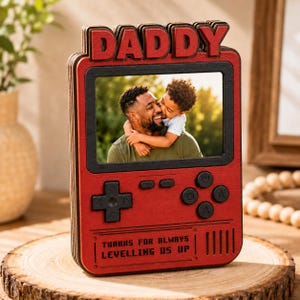 May include: A red wooden photo frame shaped like a retro game console, with the word "DADDY" in red letters at the top. The frame holds a photo of a man and a child. The text "THANKS FOR ALWAYS LEVELLING US UP" is at the bottom.