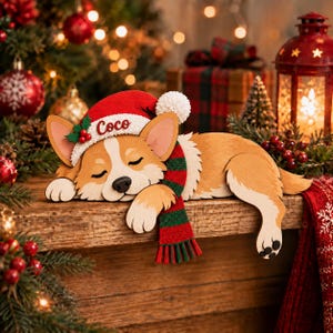 May include: A wooden cutout of a sleeping Corgi dog wearing a red Santa hat with a white pom-pom and a green and red striped scarf. The hat features the name Coco in red script and a small sprig of holly with red berries on the side.