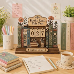 Może przedstawiać: Drewniany stojak na biurko z napisem Books Read This Year, licznikiem z klapkami wskazującym 027 oraz podstawą z napisem Sophia's Library. Element zawiera rzeźbione akcenty kwiatowe, malowane grzbiety książek i mały wazon, otoczony notatnikami i długopisami.