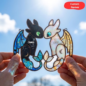 May include: A stained glass suncatcher featuring two dragon figures facing each other. The left dragon is black with blue wing accents and the name James. The right dragon is white with yellow wing accents and the name Anna. Both have green eyes.