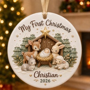 Puede incluir: Un adorno navideño redondo de cerámica blanca con una cinta dorada. El diseño muestra un pesebre con un bebé, un ciervo, un conejo y un cordero bajo una estrella dorada. El texto dice My First Christmas, Christian y 2026.